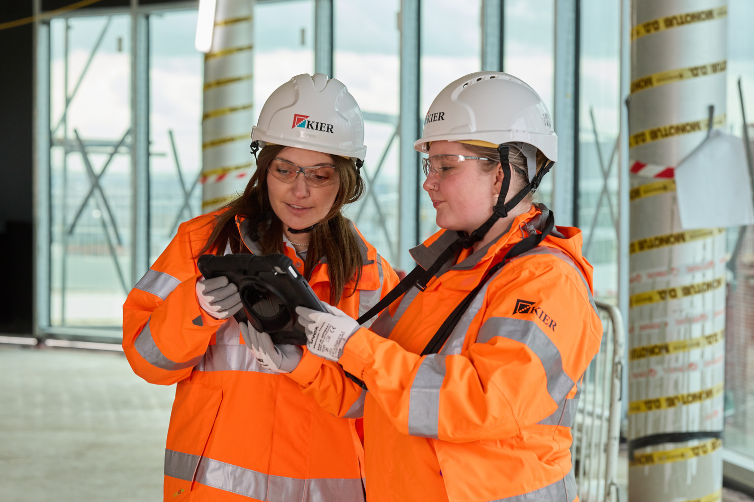 Two women in orange hi-vis jackets and white hard hats stood on a construction site looking at information on a tablet.