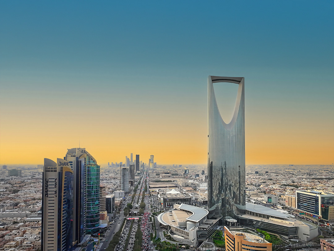 A bird's eye view of Riyadh with buildings on wither side of the street. One tall building is on the right. It is the evening with a light blue and orange sky and the sun has already set. There is a electric teal and light green neon oval over the photo.