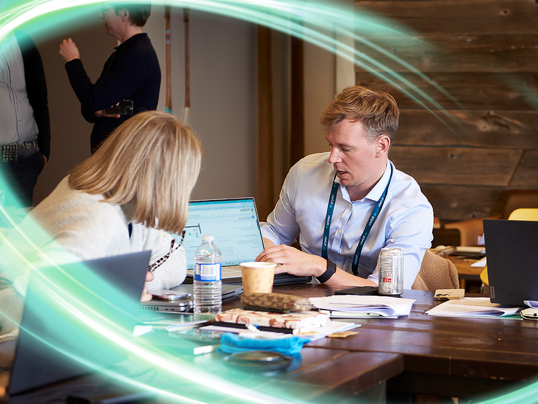 Two employees are looking at tehe same computer screen that is facing the camera and blurred out. They have coffee and water in front of them with papers on the wooden table. There is a electric teal and light green neon oval over the photo.