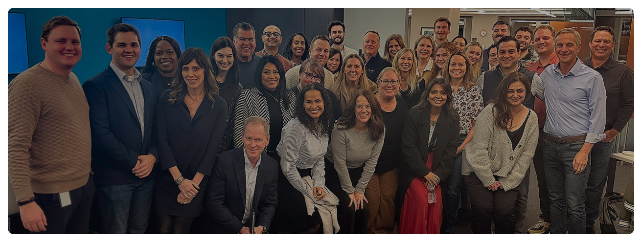 A large group of employees standing next to each other smiling in stacked lines in an office with a blue wall behind them.