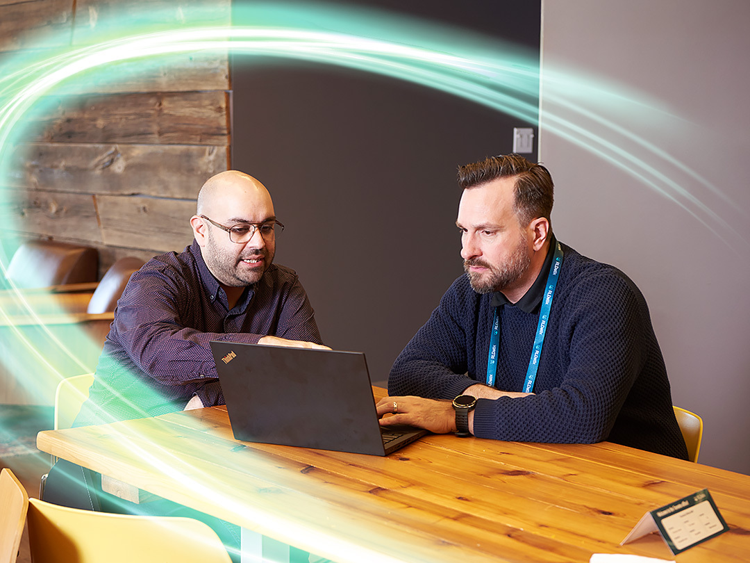 Two employees are working at a computer on an oak wood table. There is a electric teal and light green neon oval over the photo.