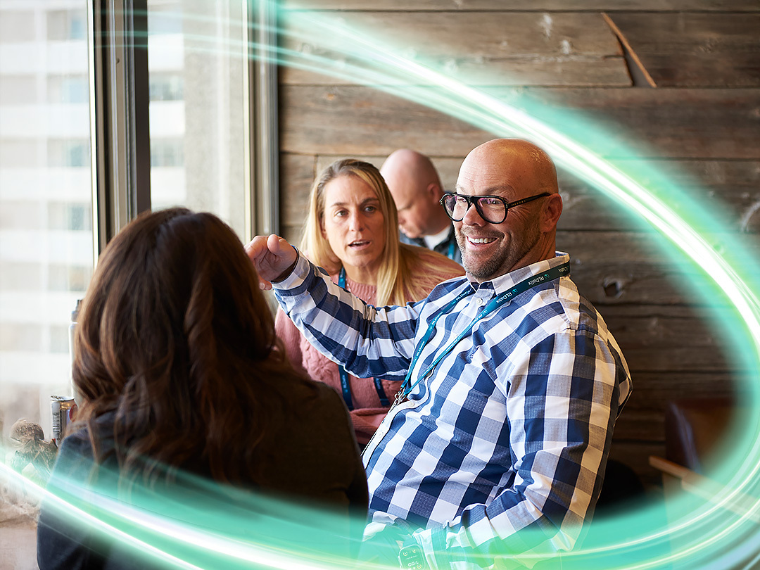 Three employees are sitting in front of a window withe a light wooden wall behind them. One is centered and smiling in a blue and white checkered shirt. He is wearing glasses. There is a electric teal and light green neon oval over the photo.