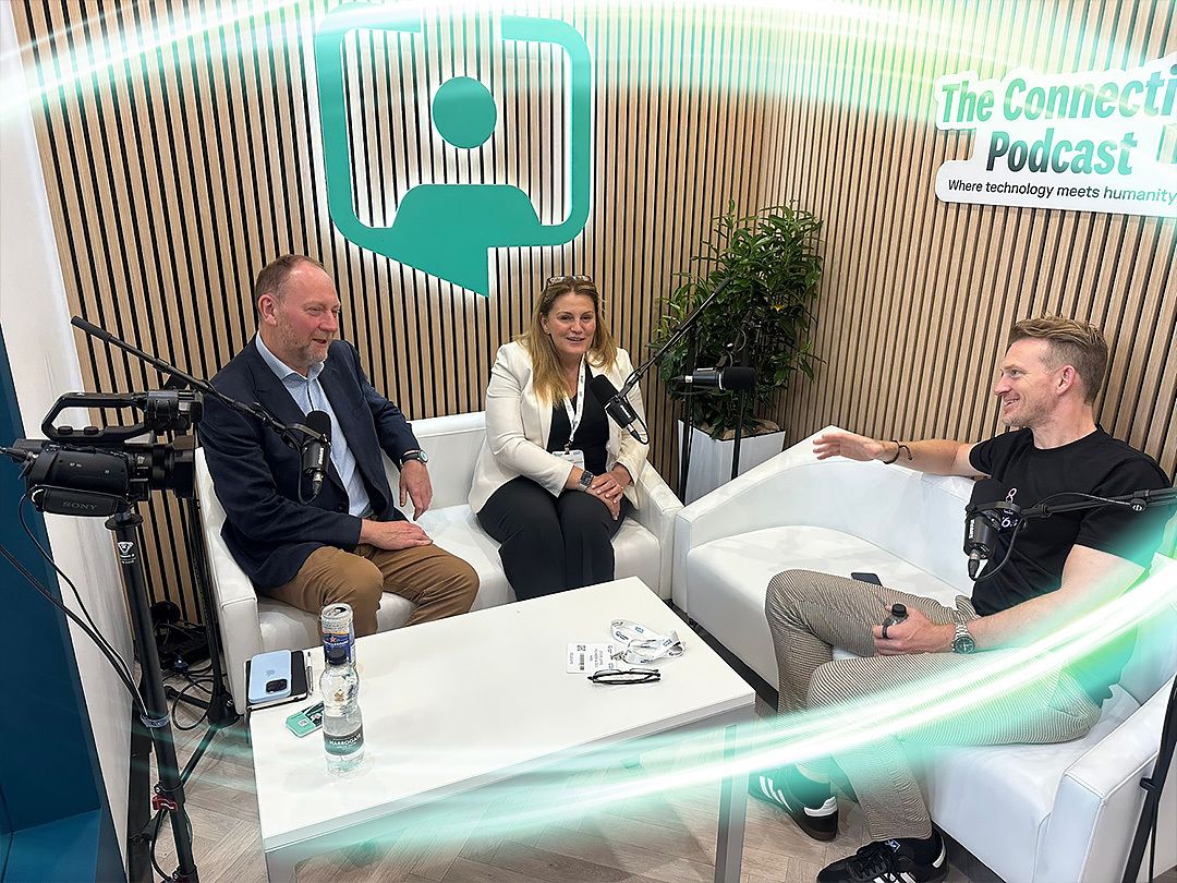 Two employees are sitting on a white couch with microphones in front of them. The RLDatix logo is behind them and there is light brown paneling on the wall. There is a electric teal and light green neon oval over the photo.