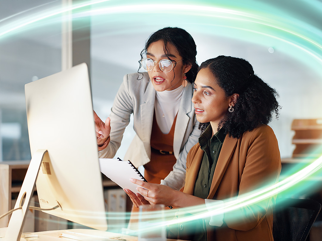 Two women in business clothes are looking at a screen and one is pointing. There is a electric teal and light green neon oval over the photo.