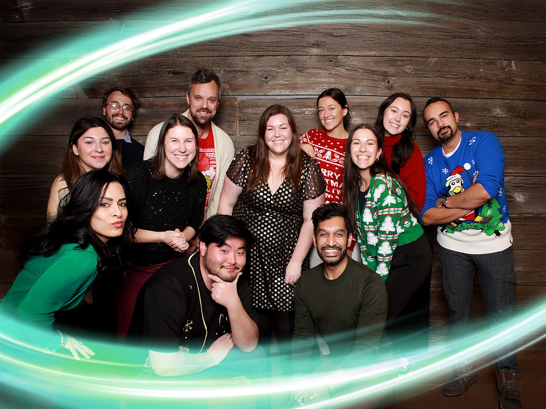 Group of employees in Christmas sweaters smiling. There is a electric teal and light green neon oval over the photo.