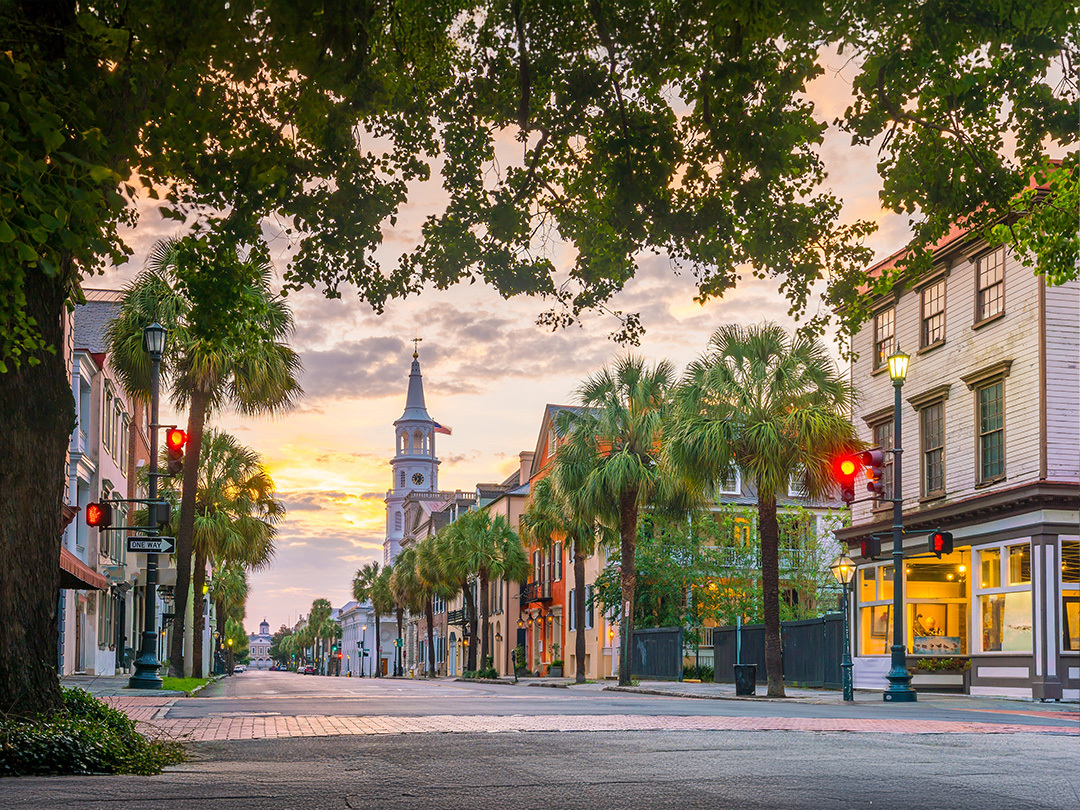 The city streets of Charleston with a tree overhanging and buildings on the right. It is the evening with a purpling and clouded sky. There is a electric teal and light green neon oval over the photo.