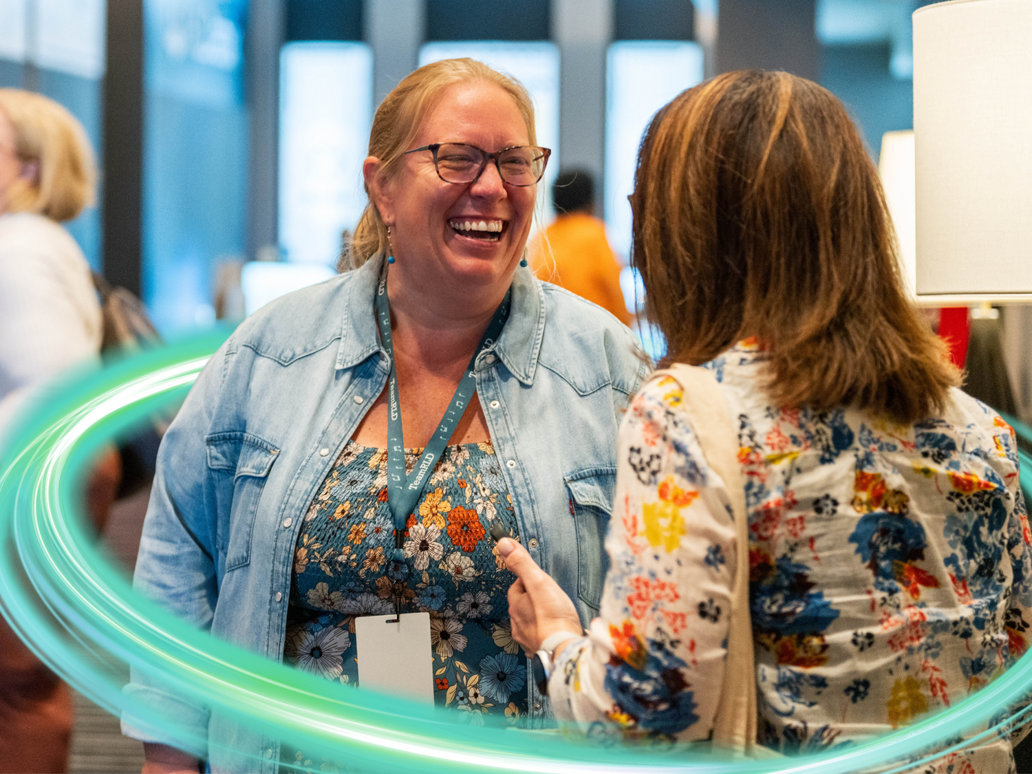 A woman speaking with another person, smiling in a long-sleeved jean shirt. There is a electric teal and light green neon oval over the photo.