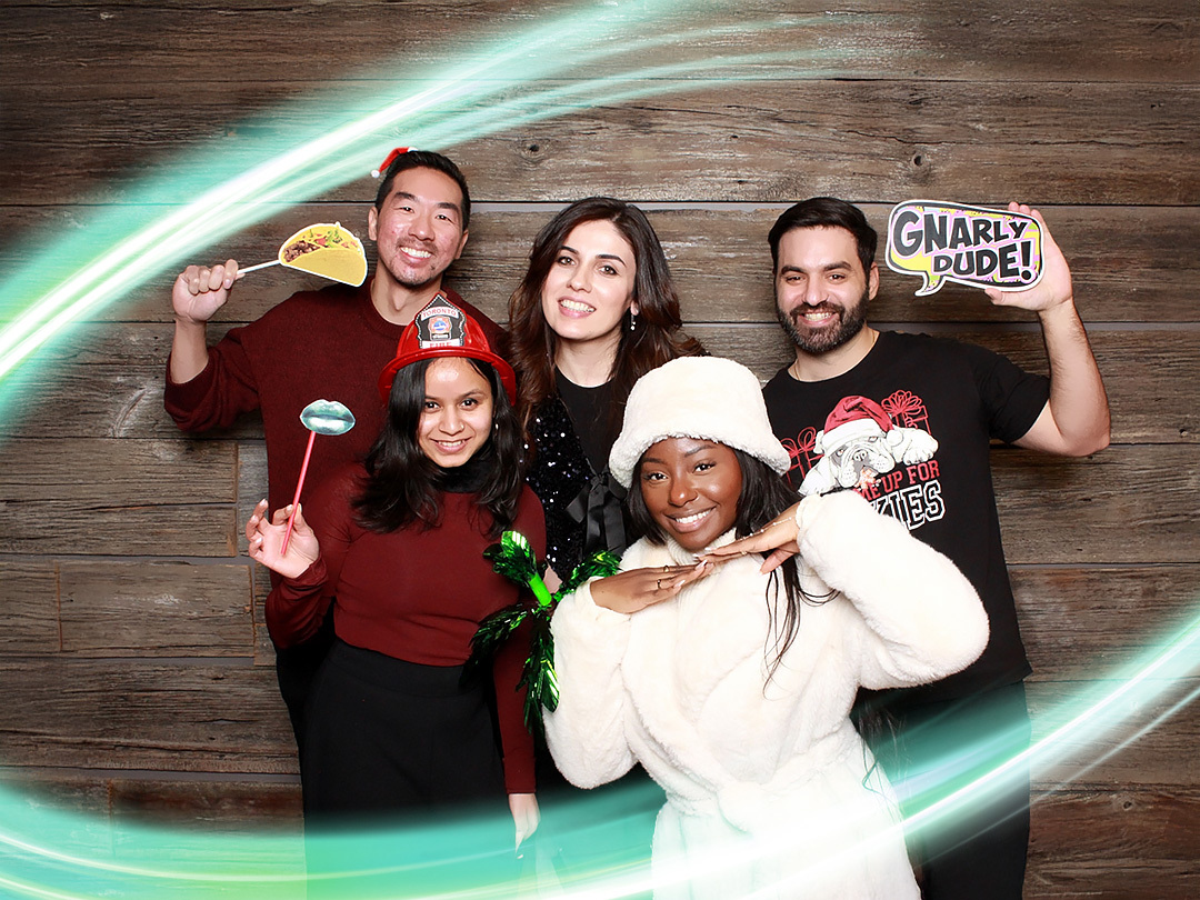 A group of 5 employees are smiling at the camera with a light wooden background. They are holding goofy signs like a taco and one person is wearing a fire helmet. There is a electric teal and light green neon oval over the photo.