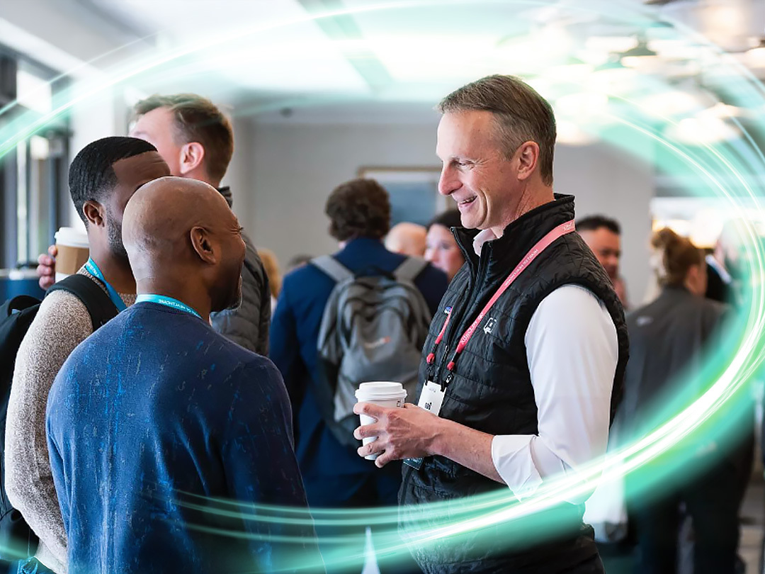 There is a man with a cup in hand and a bubble vest smiling at another person he is talking to. There are people behind them standing. There is a electric teal and light green neon oval over the photo.