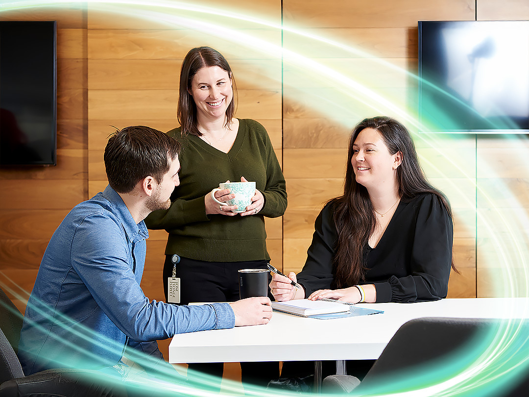 Three employees smiling in close proximity with two sitting and the third standing next to them with a coffee cup in hand.  There is a electric teal and light green neon oval over the photo.