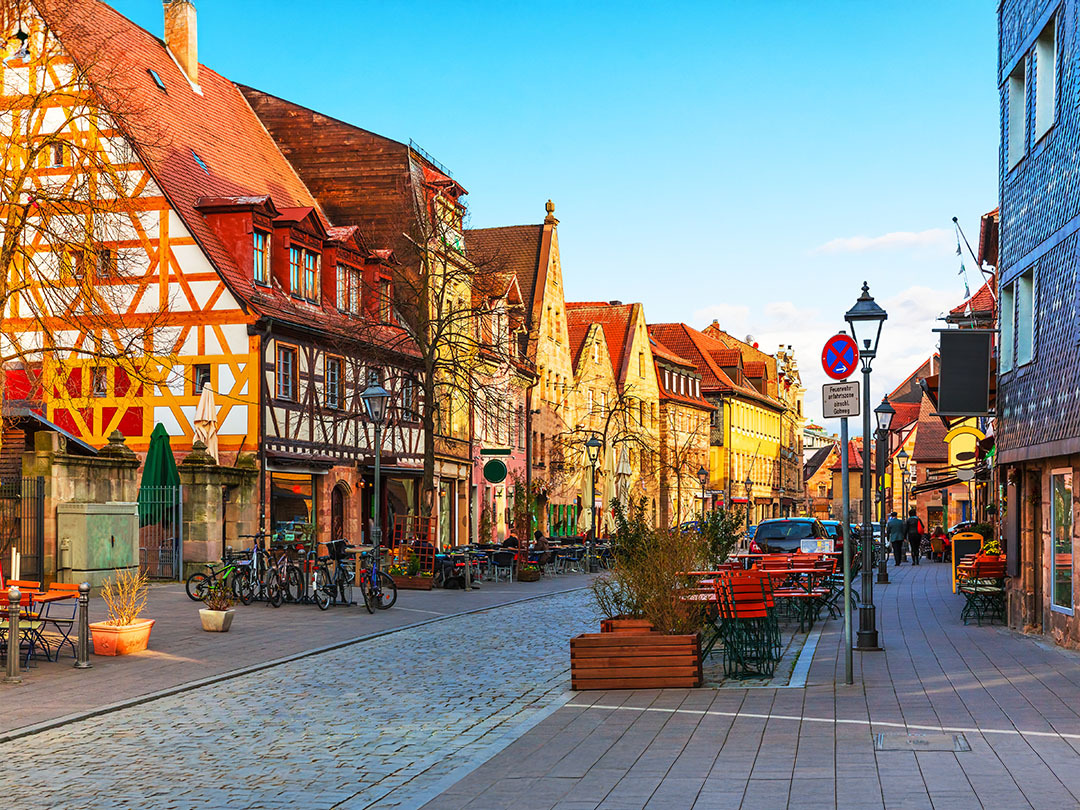 The city street of Frankfurt with older German architecture on the shorter buildings on the left. There are red flowers in front of the photo. It is the evening with a light blue colored sky. There is a electric teal and light green neon oval over the photo.