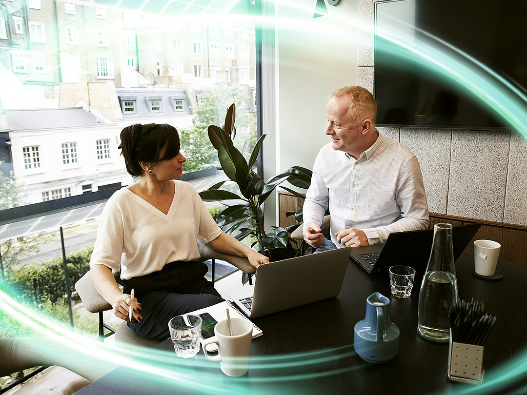 Two people are talking to each other in a room with a large window taking up the back wall and a tv mounted to the other wall. There is a plant behind them and a computer in front of them. There is a electric teal and light green neon oval over the photo.