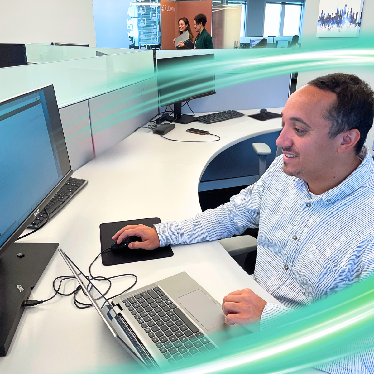A man sitting at a desk, smiling in a long-sleeved shirt. There is a electric teal and light green neon oval over the photo.