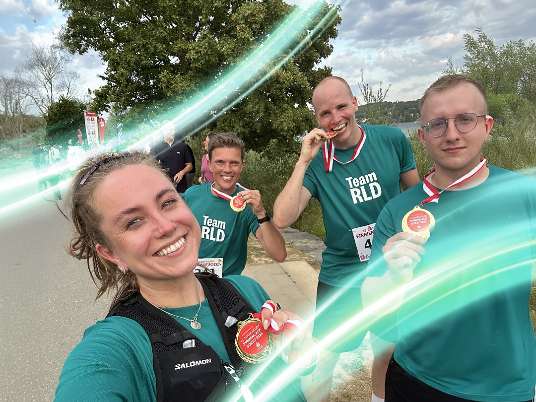 A group of four people are outside after a run. They are wearing dark green TeamRLD shirts and they have medals. There is a electric teal and light green neon oval over the photo.
