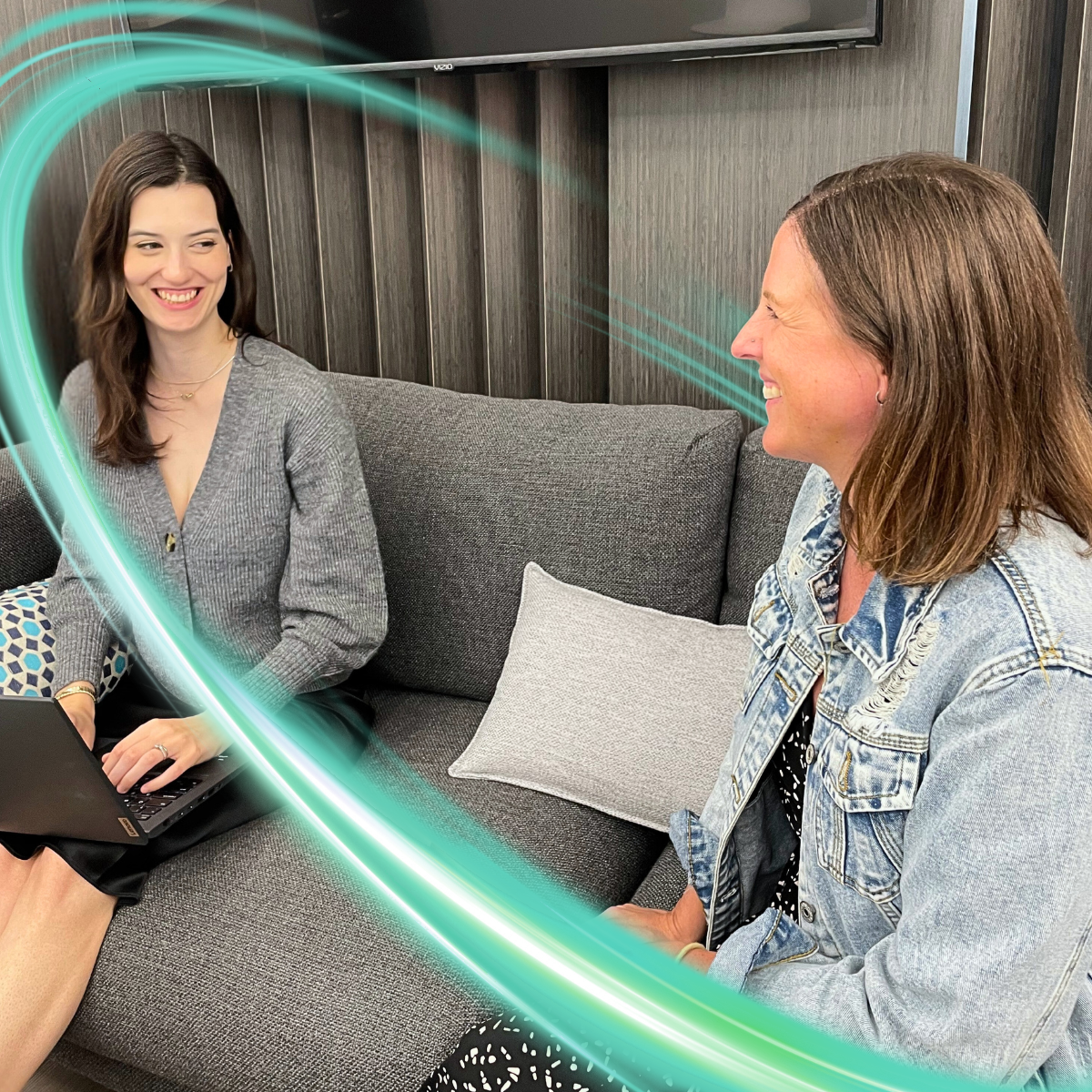 Two women are sitting on a couch looking at each other smiling. One has a computer on her lap. There is a electric teal and light green neon oval over the photo.