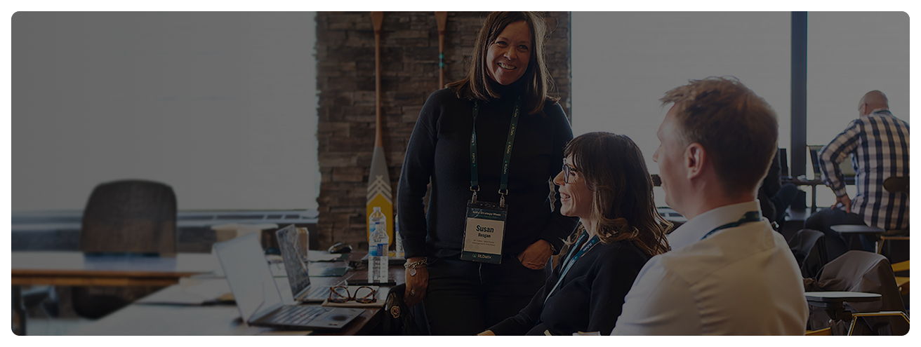 Three employees are smiling. One is standing while looking at the two sitting in front of two computers that are side by side on a large l-shaped wooden desk. There are two paddles mounted on a 1 meter wide brick wall behind them in between two large windows.