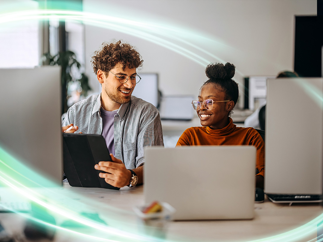 Two people are sitting next to eachother smiling. The man is looking at the woman while she is looking at his ipad. She has a computer in front of her. There is a electric teal and light green neon oval over the photo.