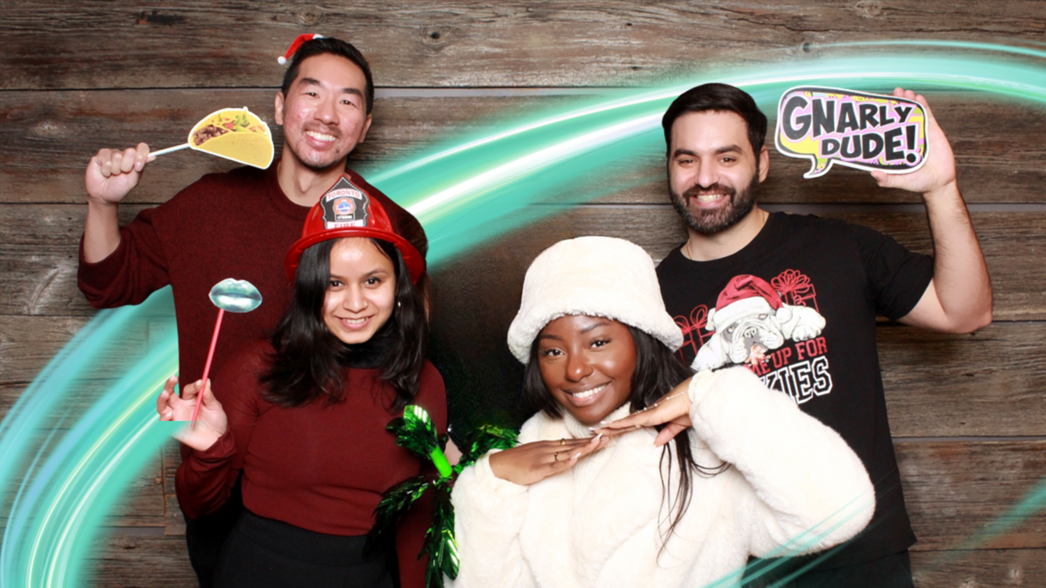 A group of 5 employees are smiling at the camera with a light wooden background. They are holding goofy signs like a taco and one person is wearing a fire helmet. There is a electric teal and light green neon oval over the photo.