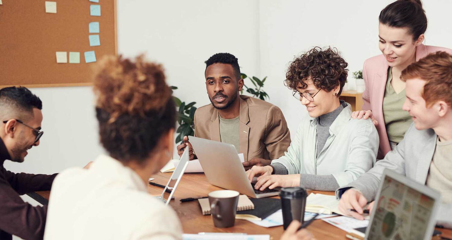 Six people sitting at a wooden table. One is smiling and looking at their computer. The other seems to be in mid-conversation.