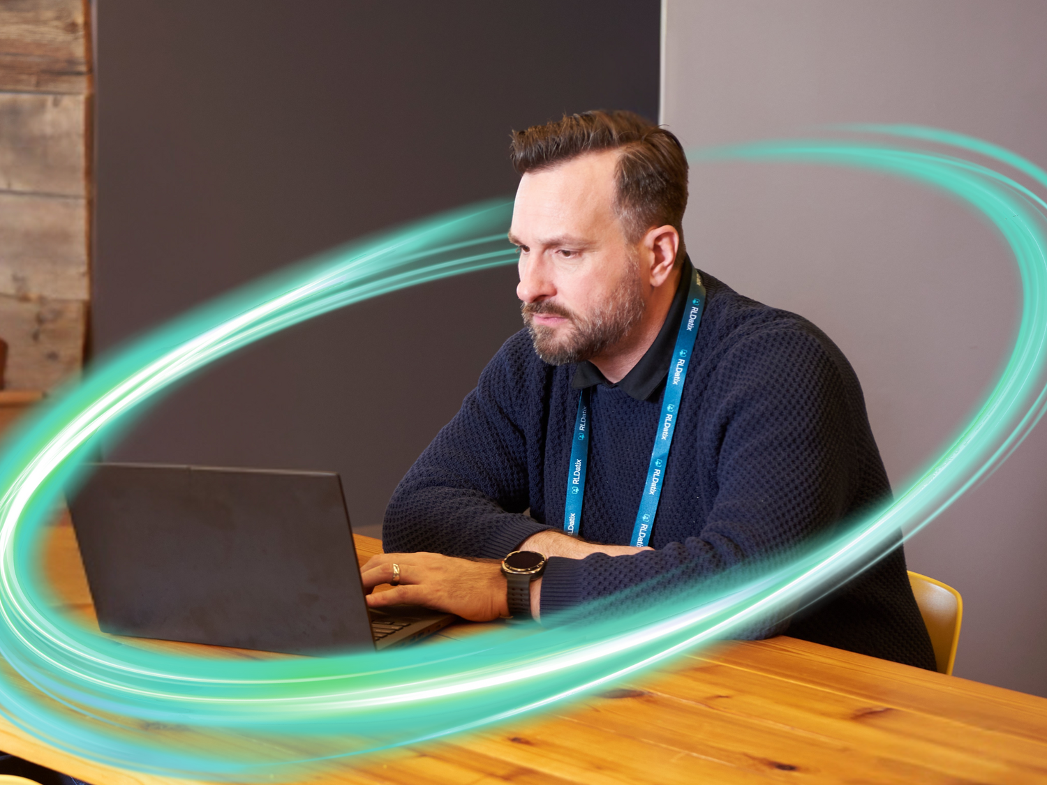 An employee is working at a computer on an oak wood table. There is a electric teal and light green neon oval over the photo.