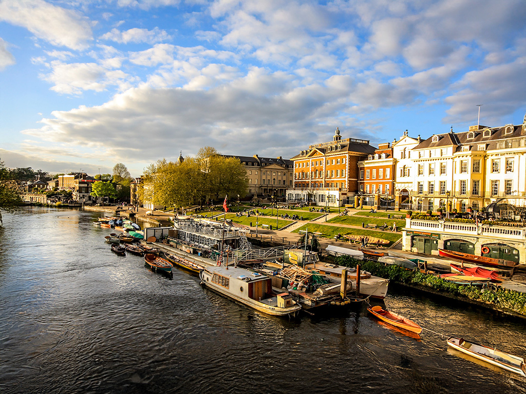 The city in the UK with shorter buildings in the back and a river in the front. It is the evening with a light blue and clouded sky. There is a electric teal and light green neon oval over the photo.